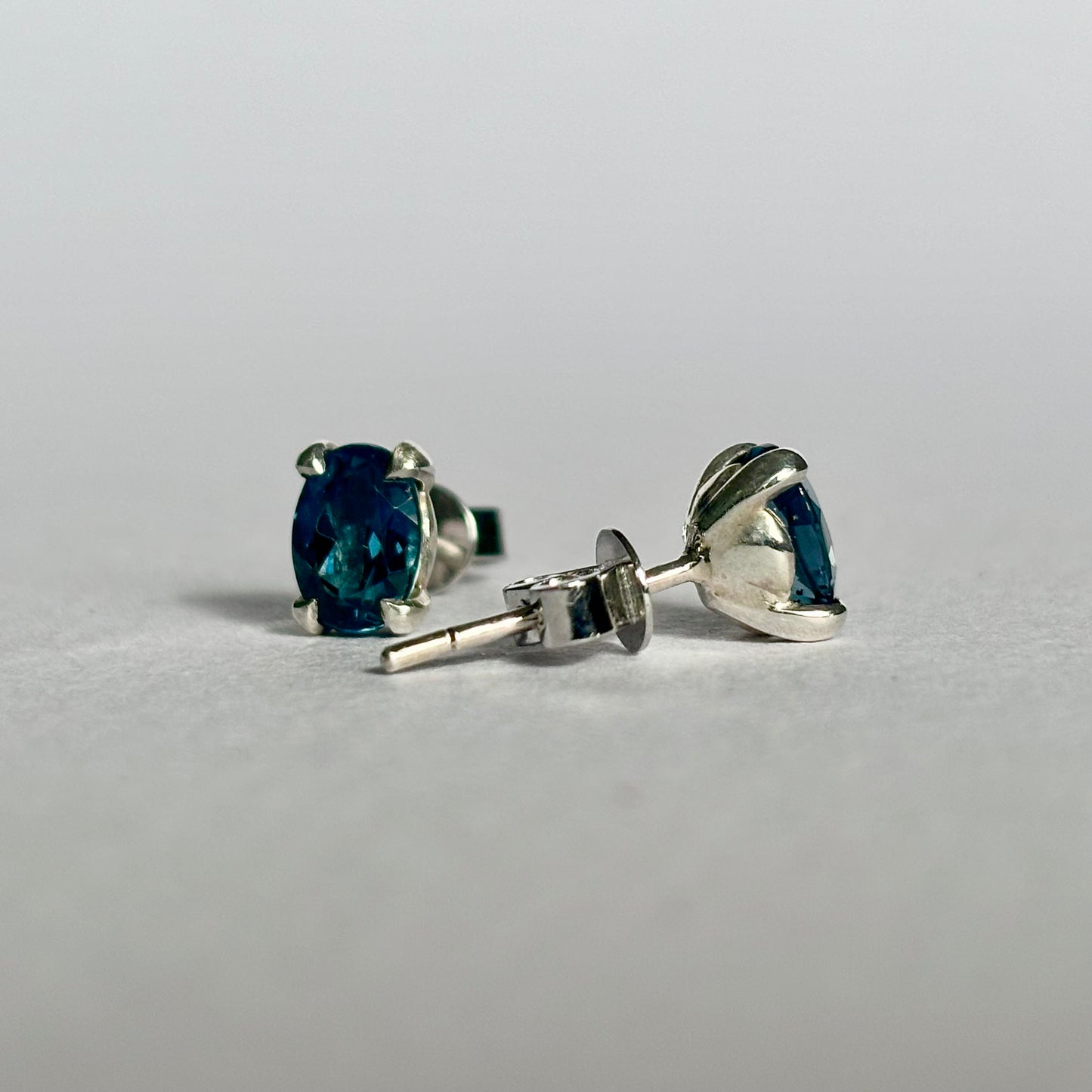 A pair of 9ct white gold stud earrings with oval-cut teal sapphires , one shown from the front and the other from the side/back, on a light gray background.